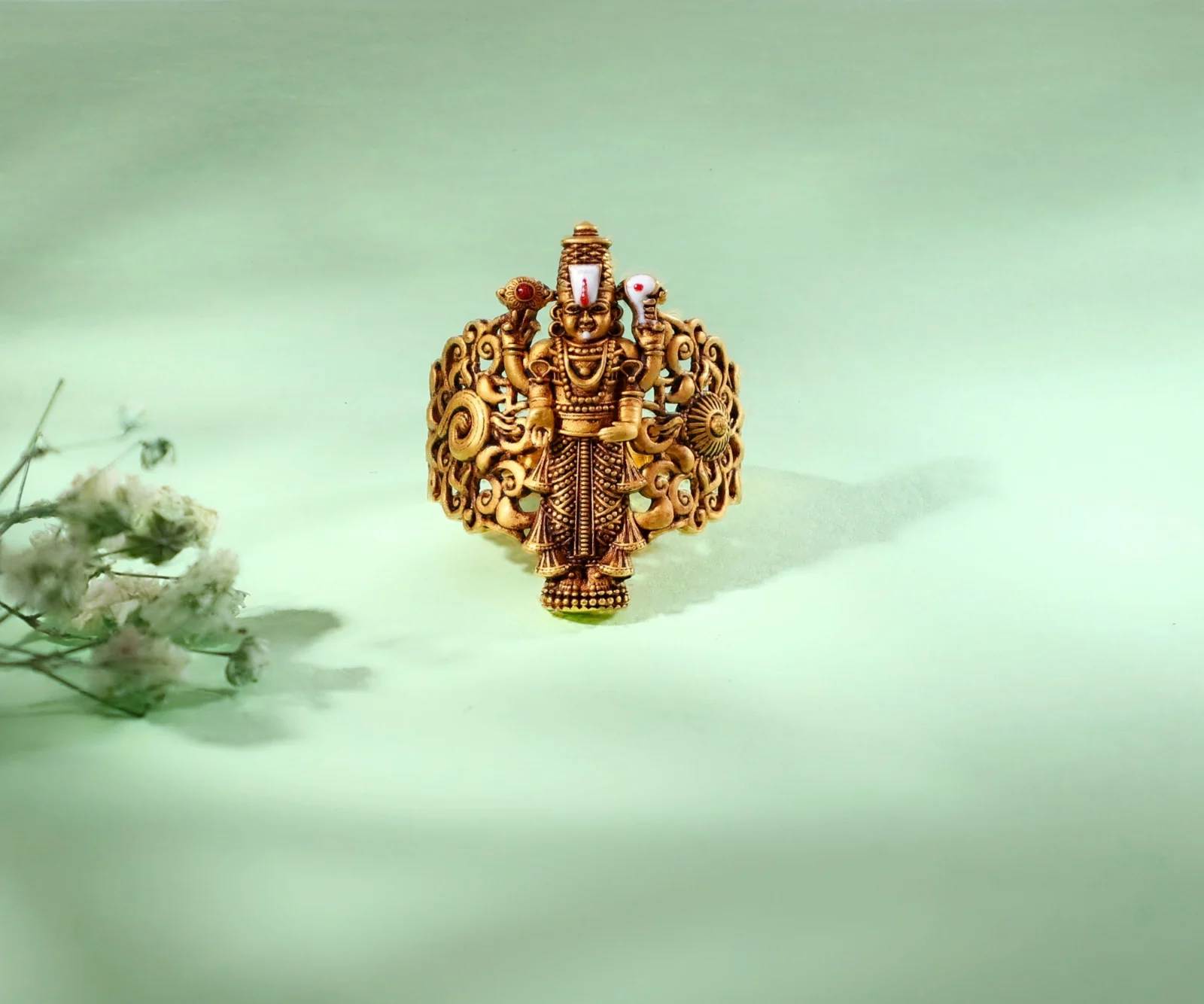 An antique-gold Balaji ring on a pale green backdrop with blurred dried flowers in the foreground
