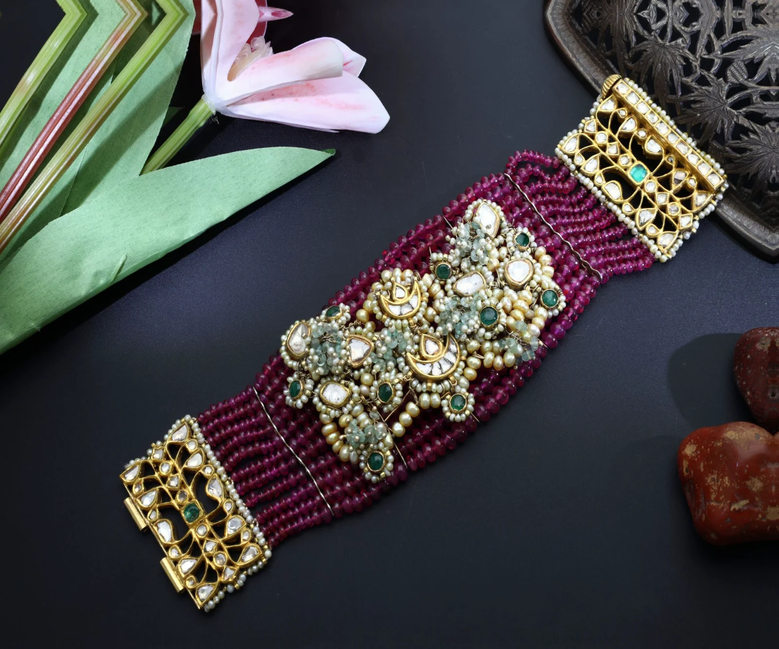 A multi-strand red-bead bracelet with kundan clasps laid flat on a dark surface with a lily and an antique tray