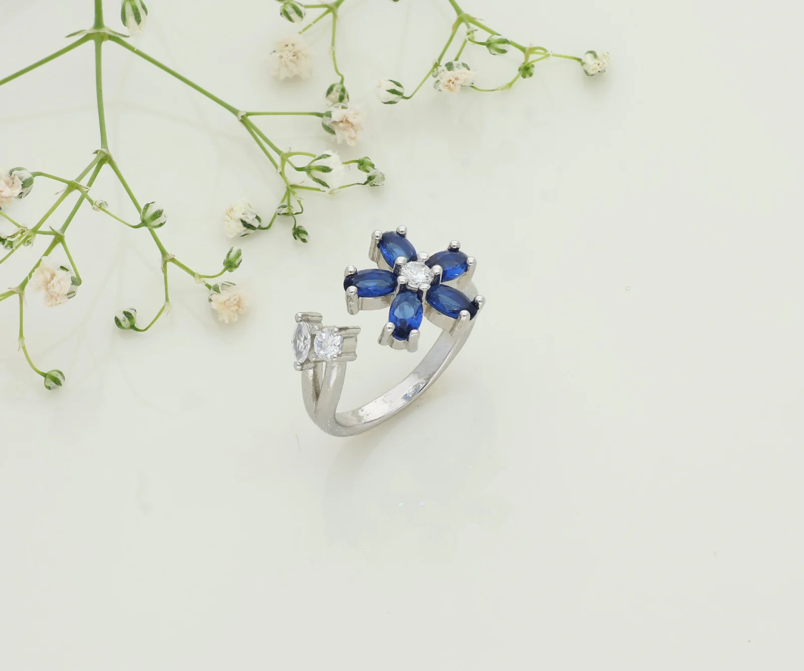 A silver open ring with a blue-stone flower on an ivory backdrop with sprigs of baby’s breath