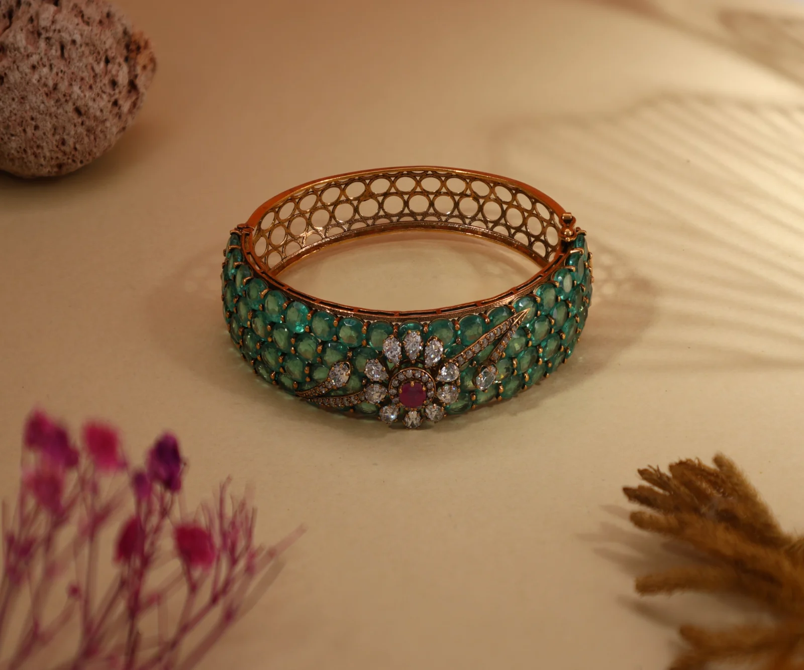 A green-stone bangle with a floral centre on a beige backdrop with leaf shadows and blurred flowers
