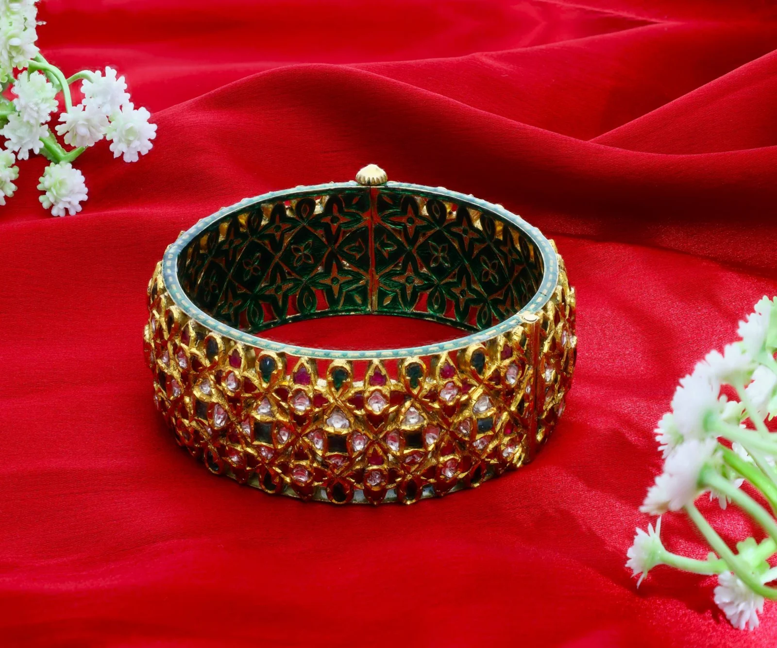 A kundan bangle with enamel work inside, placed on folded red silk with white flowers