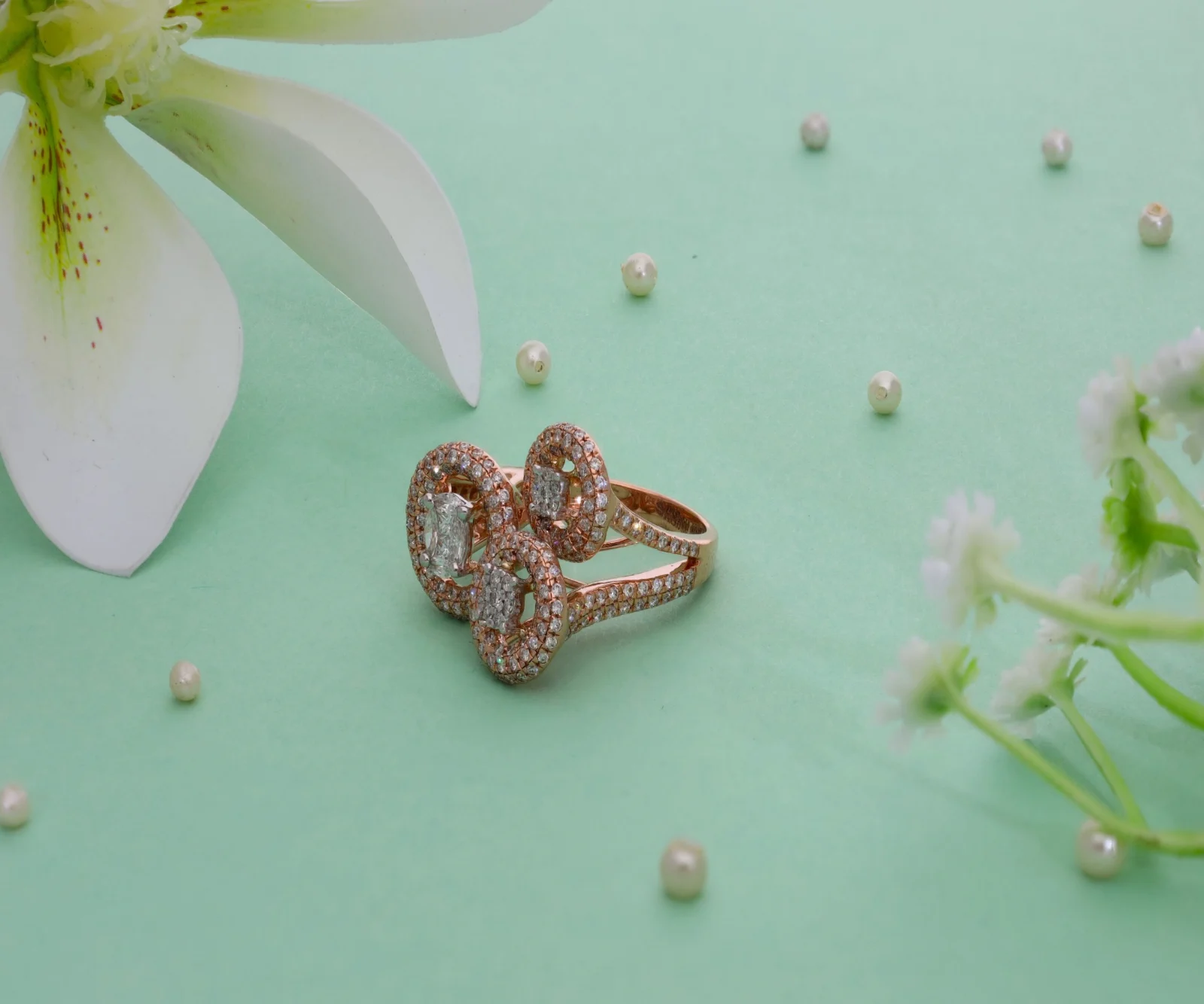 A rose-gold cocktail ring on a mint backdrop with a white lily and scattered pearls